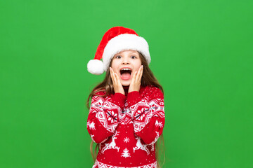 A little girl shows delight at a Christmas gift. A child with long hair in a reindeer sweater and Santa Claus hat on a green isolated background.