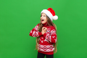 A little girl shows delight at a Christmas gift. A child with long hair in a reindeer sweater and Santa Claus hat on a green isolated background.