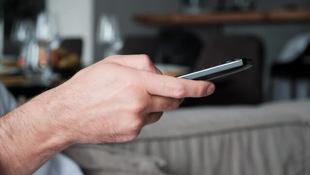 Close up of man's hand selects internet tv channels with remote control in modern apartments. Male controls TV using a modern remote control. A man watches smart TV and uses black remote control. 