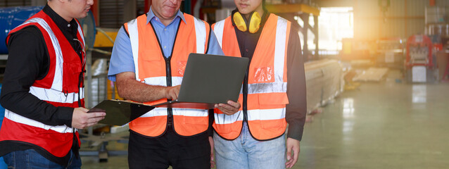 A manager using an empty laptop schematically with a team of multi-ethnic male engineers in safety clothing stands and talks in the factory.