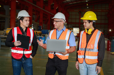 A manager using an empty laptop schematically with a team of multi-ethnic male engineers in safety clothing stands and talks in the factory.