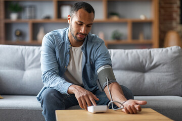 Black Man Sitting On Couch, Checking Blood Pressure With Upper Arm Monitor