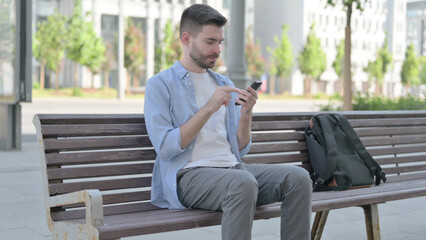 Young Man Browsing Internet on Smartphone while Sitting on Bench