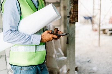 Construction engineers wear safety hardhat and in vest hold a blueprint and use a mobile phone on...