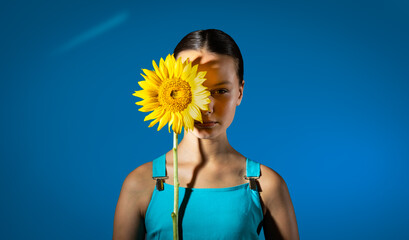 Fashion portrait of beautiful preteen serious girl with yellow sunflower in hands against blue background