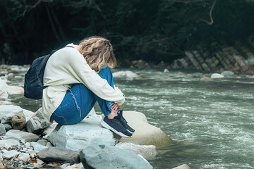 frustrated, tired tourist girl sits alone on the bank of a mountain river on the rocks, she clasped her legs in her hands and laid her head on them during a hike in nature