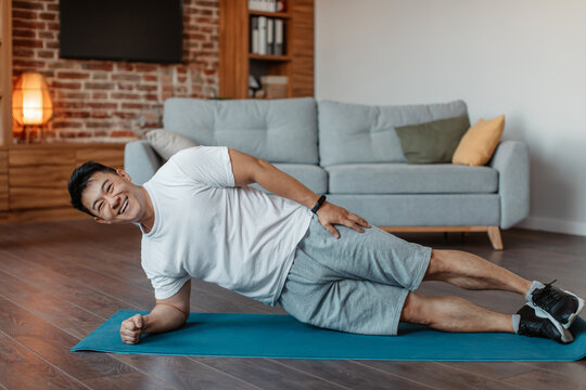 Happy Asian Mature Man In Sportswear Doing Side Plank On Floor Mat In Living Room Interior, Copy Space