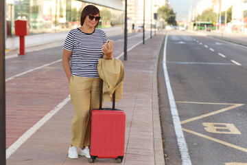 senior woman in sunglasses with red suitcase is using smartphone outdoors by the airport or railway station waits for taxi. Female ready to travel with luggage in the modern city.