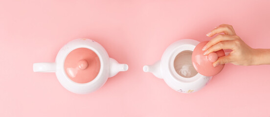 Banner. Two white porcelain teapots with a pink lid on a pink background. Female hand removes the lid from the kettle. Flat lay. Copy space. Concept of Breast Cancer Awareness Month