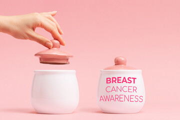 Two white porcelain pots with a pink lid on a pink background. Female hand removes the lid from the left side jar. Concept of Breast Cancer Awareness Month