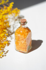 Transparent bottle filled with yellow bath sea salt with wild flowers on white background. Beauty treatment for spa and wellness. Skincare natural cosmetic concept for body care.