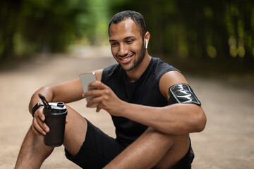 Happy african american sportsman drinking protein cocktail, using smartphone