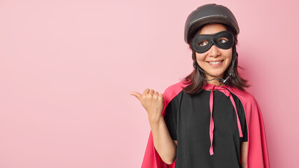 Positive Asian woman wears superhero costume with cloak prepares for party points thumb on left shows blank space for your promotional content has good mood isolated over pink background. Look at this