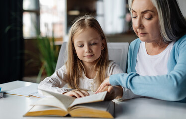 Concentrated european little girl and mature woman reading book on table in living room interior