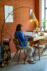 Woman works on laptop while sitting by wooden table at cozy living space at home. Concept of remote creative work and freelance