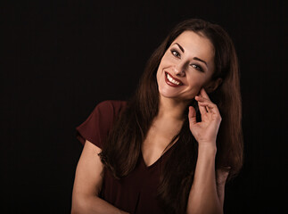 Beautiful young business toothy smiling woman thinking and looking happy in burgundy blouse and with red lipstick on dark shadow black background with empty copy space. Portrait