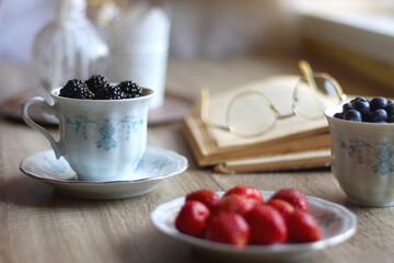 Vintage tea set filled with blueberries, blackberries and starwberries, open book, reading glasses and decorative candles on the table. Selective focus.