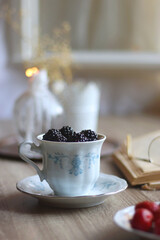 Vintage tea set filled with blueberries, blackberries and starwberries, open book, reading glasses and decorative candles on the table. Selective focus.