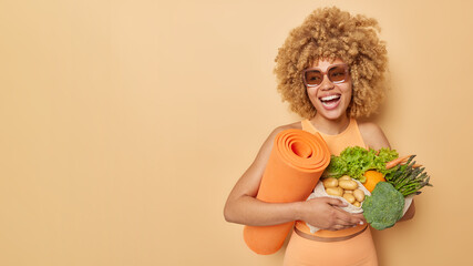 Cheerful sporty woman with curly hair dressed in sportswear laughs gladfully carries fitness mat...