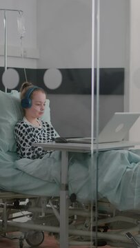 vertical video: Alone sick girl patient with oxygen nasal tube relaxing in bed wearing headphones while watching entertainment movie on laptop computer during medical examination. Girl recovery after