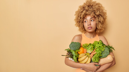 Horizontal shot of pensive curly haired woman looks away being deep in thoughts carries variety of fresh vegetables and fruits keeps to healthy diet isolated isolated over beige studio background