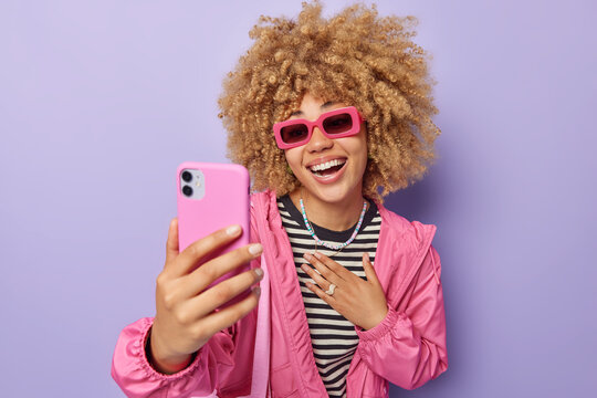 Positive curly haired young European woman wears trendy sunglasses striped jumper and jacket takes selfie makes video call has happy conversation with best friend isolated over purple background.