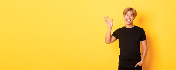 Portrait of handsome friendly asian guy in black outfit, waving hand to say hello and smiling,...