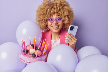 Positive curly haired woman smiles happily holds smartphone invites friends to birthday party holds...
