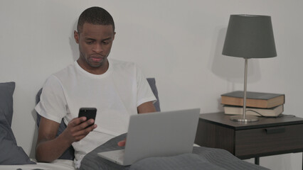 African Man with Laptop using Smartphone, Sitting in Bed