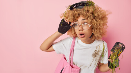 Shocked woman with curly hair feels impressed wears protective helmet spectacles and gloves holds pedal from bicycle looks aside holds breath from amazement isolated over pink background copy space