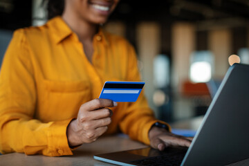 Black businesswoman holding credit card and using laptop for online shopping, sitting at workplace in office
