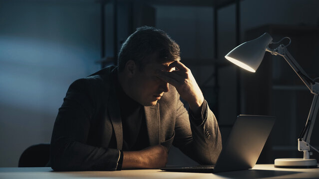 Tired man. Hard work. Remote job. Exhausted business guy suffering headache looking laptop sitting desk dark room interior.