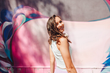 a young excited woman in a top and skirt, wearing headphones, listening to music, walking, dancing with her hands up in the open air against a pink wall.