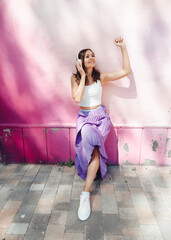 A young happy  woman dressed in a white top and purple skirt, headphones, listening to music, dancing on a city street near the wall of a building. The concept of the lifestyle of urban youth People.
