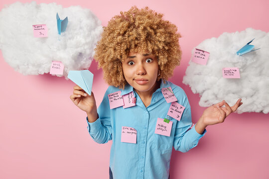 Puzzled shocked woman doesnt know from what to start has many tasks to finish holds paper handmade plane wears blue shirt makes notes on stickers poses around white clouds against pink background