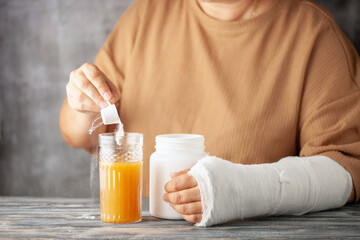 Woman with a cast on her arm pours collagen into a glass of juice on the  grey background. Girl has a broken hand wearing