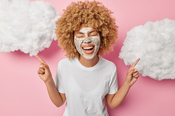 Beauty treatments concept. Joyful woman with curly hair applies nourishing clay mask smiles broadly dressed in casual white t shirt points index fingers above stands over pink backgroud white clouds