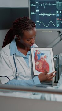 Vertical video: Medic pointing at tablet with cardiology image on display, explaining diagnosis to young patient in bed. Physician using gadget with heart organ image to show cardiovascular figure