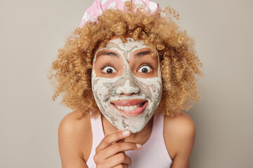 Young curly haired woman bites lips stares wondered applies clay mask to reduce fine lines wears bathhat and t shirt undergoes skin care procedures isolated over grey background. Wellness concept