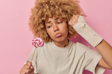 Upset curly haired young woman looks sadly at sweet lollipop on stick wants to eat candy wears beige t shirt elastic bandage on arm after injury or sprain has bad mood isolated over pink background.