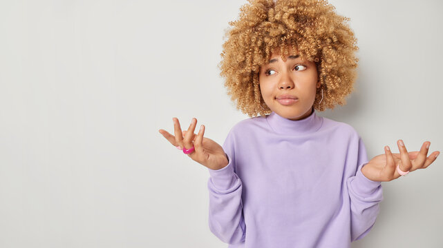Horizontal shot of confused curly haired woman spreads palms has no clue cant decide stands puzzled upset with lack of choices has no idea what to do wears purple pullover isolated on grey background