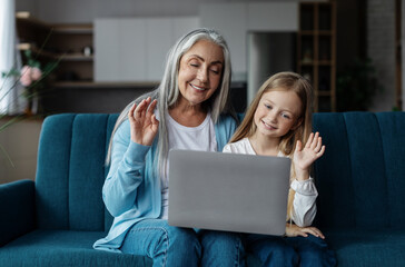 Happy european small girl and mature woman look at laptop, waving hand in living room interior