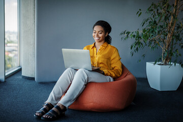 Positive black female manager working on laptop computer in office, sitting on pouf beanbag chair, free space