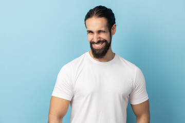 Portrait of cheerful man with beard wearing white T-shirt being in good mood, smiling broadly and winking at camera with toothy smile. Indoor studio shot isolated on blue background.