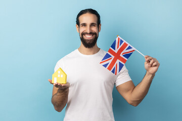 Portrait of man with beard wearing white T-shirt holding british flag and paper house, dreaming to...