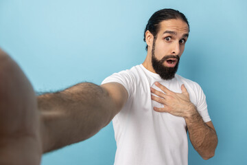 Portrait of scared shocked man with beard wearing white T-shirt looking at camera open mouth, keeps hand on chest, POV, point of view of photo. Indoor studio shot isolated on blue background.