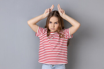 Portrait of aggressive bully little girl wearing striped T-shirt showing bull horns gesture over...