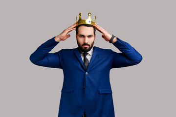 Serious independent bearded man wearing golden crown, looking with arrogance and confidence, privileged status, wearing official style suit. Indoor studio shot isolated on gray background.