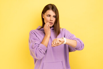 Displeased nervous woman biting finger nails and looking at her wrist clock with anxious, worried about deadline, wearing purple hoodie. Indoor studio shot isolated on yellow background.