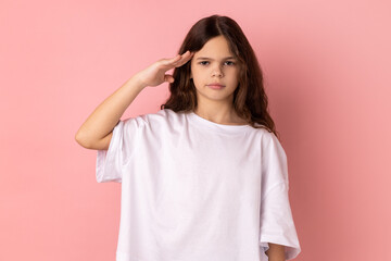 Portrait of serious little girl wearing white T-shirt giving salute, obediently listening to commander order with attentive confident face. Indoor studio shot isolated on pink background.